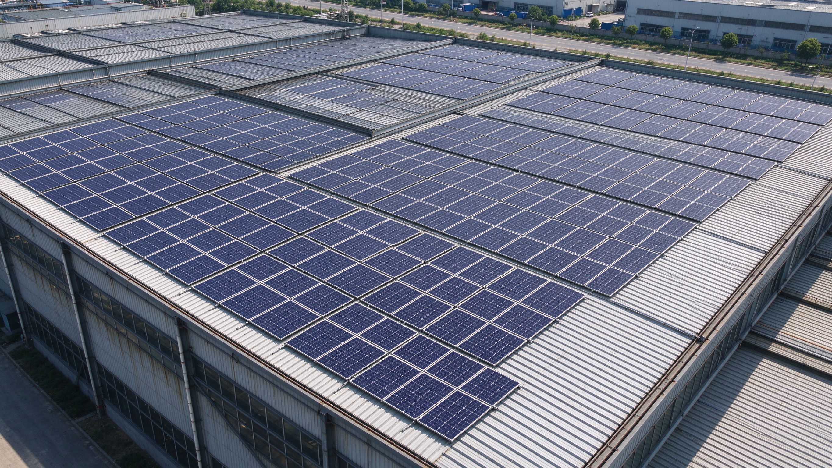 Aerial view of a steel structure industrial building with rows of solar panels mounted on the sloped metal roof (illustrative concept image)